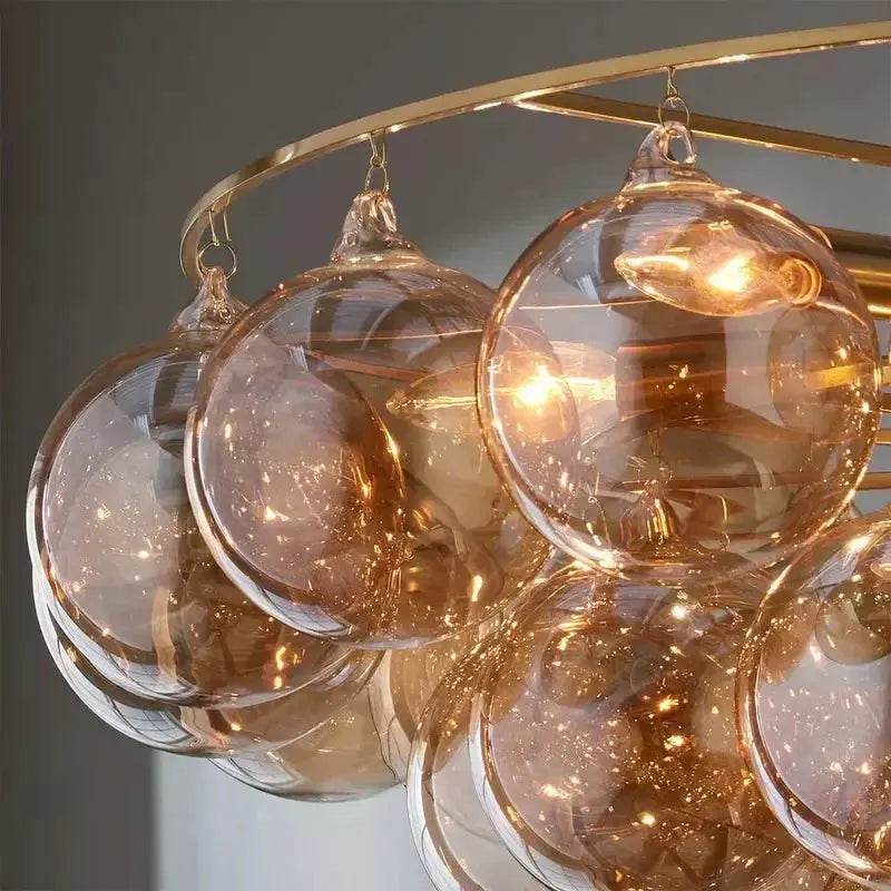 A close-up of a modern chandelier featuring multiple large, clear glass hanging spheres with warm amber light glowing inside, suspended from a curved gold-toned metal frame.