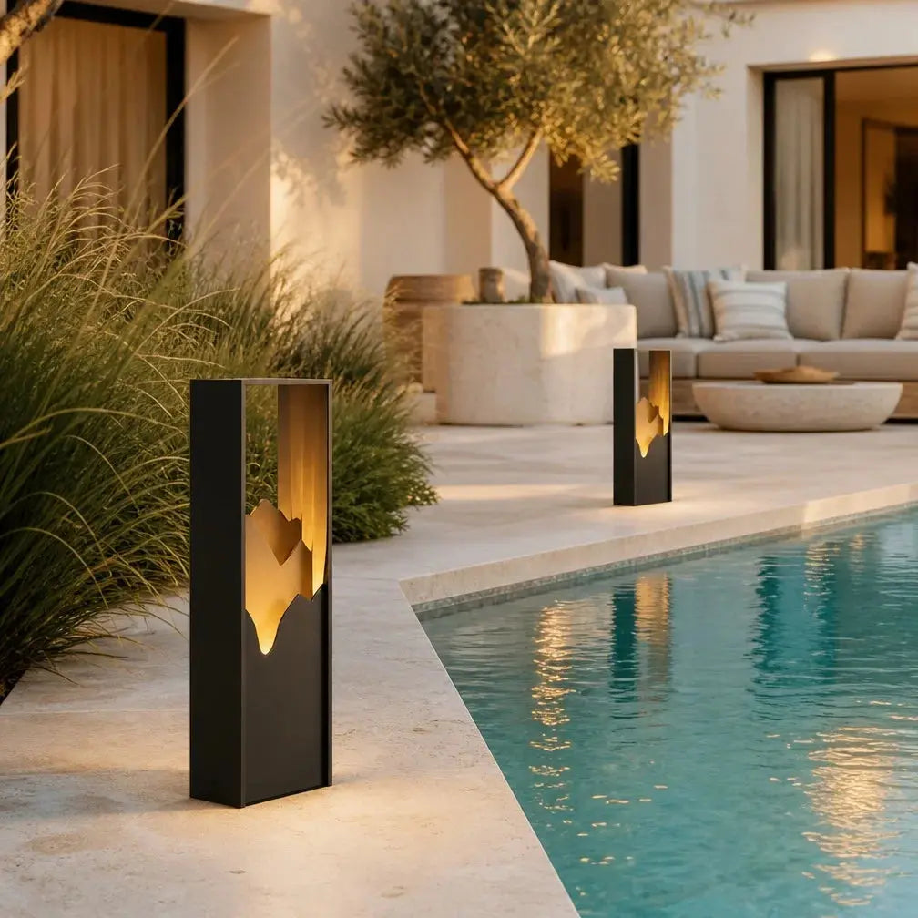 Two modern rectangular outdoor bollard lights with a matte black finish and warm glowing organic cutout panels, placed beside a swimming pool in a luxurious patio setting.