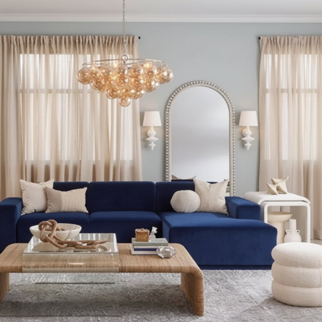 A styled living room featuring a prominent deep navy blue velvet sectional sofa, styled with beige and white pillows, a glass-top coffee table, a large arched mirror, and a glowing chandelier.