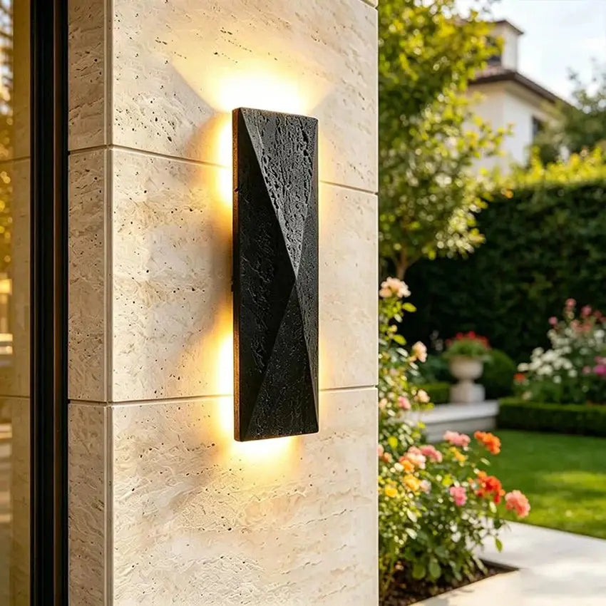 A modern rectangular black outdoor wall light with a textured surface and warm glowing LED strips, mounted on a beige stone pillar in a landscaped garden setting.